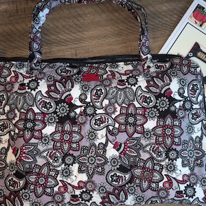 May include: A rectangular quilted tote bag featuring a repeating floral and paisley pattern in shades of black, white, grey, and deep red. The bag has two matching fabric handles, a black zip closure, and a small rectangular red brand tag.