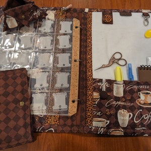 May include: A brown fabric organizer with a zipper closure and multiple pockets. The organizer has a coffee theme with images of coffee cups and beans. It includes a clear plastic sheet with slots for thread spools, a ruler, and a small notebook.