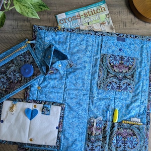 May include: A blue quilted project bag with multiple pockets and compartments. The bag has a zippered mesh pocket, a large pocket with a flap, and several smaller pockets for storing sewing supplies. The bag is decorated with a floral pattern.
