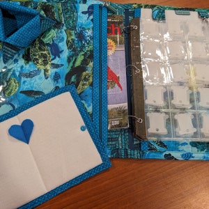 May include: A blue fabric binder with a clear plastic insert for storing embroidery floss. The binder has a sea turtle design and a small blue heart on the front cover. The binder is open to reveal a magazine and a clear plastic sheet with multiple compartments for storing floss.