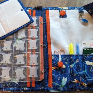 May include: A blue and orange fabric craft organizer with a dragonfly pattern. It features a clear plastic floss holder, a small notebook, and pockets for scissors, pens, and other tools. The organizer is open, revealing its contents.