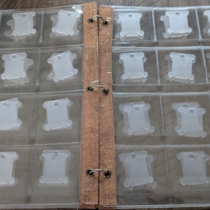 May include: A clear plastic storage binder with twenty-four individual pockets designed for organizing embroidery floss bobbins. The binder features a central spine made of light brown cork material with three metal rings holding the pages together.