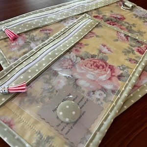 May include: Three clear zippered pouches with green and white polka dot fabric and a floral print lining. The pouches are stacked on top of each other. The top pouch has a silver ring attached to it. The bottom pouch has a small card inside that says "NeaMinder Button) This contains a magnet and is designed to keep out of reach of children and pets."