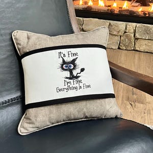 May include: A beige pillow with black trim features an embroidered black cat with the text "It's Fine, I'm Fine, Everything Is Fine."
