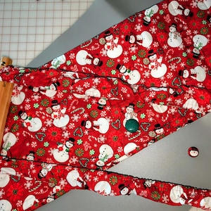 Puede incluir: Tela roja con un patrón repetido de muñecos de nieve blancos, bastones de caramelo y copos de nieve. La tela está doblada por la mitad y tiene una varilla de madera unida a ella.