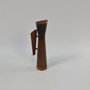May include: A copper and wood vase with a textured design. The vase has a handle on the side and a black interior.