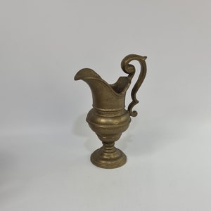 May include: A small, gold-colored metal pitcher with a decorative handle. The pitcher has a round base and a slightly flared top.