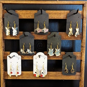 May include: A display of 10 pairs of earrings on black and white card stock. The earrings feature various designs including bats, skulls, and animals. The earrings are displayed on a wooden shelf.