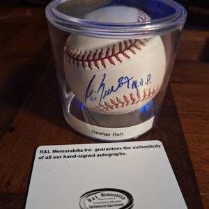 Op de afbeelding: Een witte honkbal gesigneerd door George Bell, tentoongesteld in een doorzichtige plastic vitrine met een blauwe lichtbasis. De vitrine staat op een houten oppervlak. De kaart onder de vitrine luidt "R&L Memorabilia Inc. garandeert de authenticiteit van al onze handgesigneerde handtekeningen." en bevat een logo met de tekst "R&L Memorabilia Rare & Limited Hand Signed Collectibles Authenticity Guaranteed."