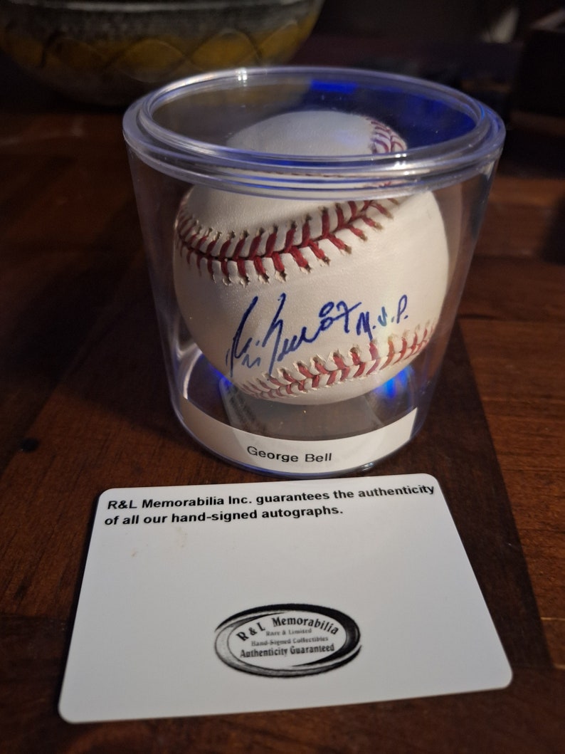 Op de afbeelding: Een witte honkbal gesigneerd door George Bell, tentoongesteld in een doorzichtige plastic vitrine met een blauwe lichtbasis. De vitrine staat op een houten oppervlak. De kaart onder de vitrine luidt "R&L Memorabilia Inc. garandeert de authenticiteit van al onze handgesigneerde handtekeningen." en bevat een logo met de tekst "R&L Memorabilia Rare & Limited Hand Signed Collectibles Authenticity Guaranteed."
