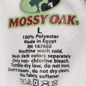 May include: White fabric tag with the Mossy Oak logo in green and red. The tag includes the text "L", "100% Polyester", "Made in Egypt", and washing instructions. The tag also has care symbols.
