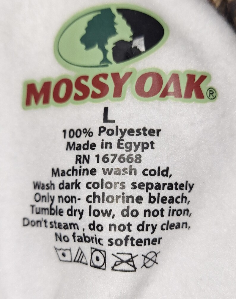 May include: White fabric tag with the Mossy Oak logo in green and red. The tag includes the text "L", "100% Polyester", "Made in Egypt", and washing instructions. The tag also has care symbols.