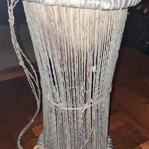 May include: A traditional African drum with a brown leather head and a woven body made of natural fibers. The drum has a rope handle and is decorated with a black band around the top and bottom.