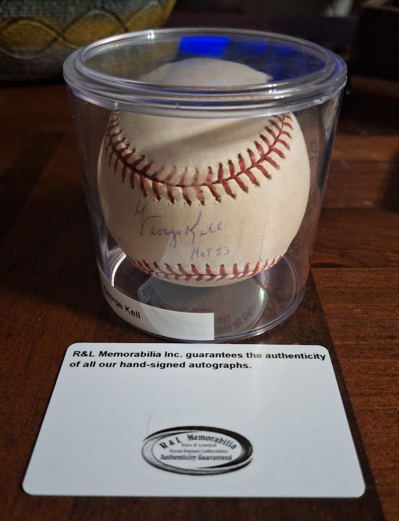 Op de afbeelding: Een witte honkbal gesigneerd door George Kell, een Hall of Fame honkballer, met de inscriptie "HOF '83". De honkbal bevindt zich in een doorzichtige plastic container met een witte kaart eronder die luidt: "R&L Memorabilia Inc. garandeert de authenticiteit van al onze handgesigneerde handtekeningen." De kaart heeft ook een rond zegel met de tekst "R&L Memorabilia Echtheidsgarantie".