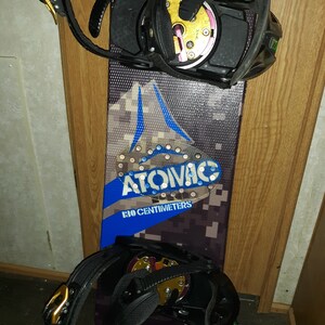 May include: A black snowboard with black and gold bindings. The snowboard has a blue and black camouflage pattern with the text "ATOIVAC 130 CENTIMETERS" printed on it.