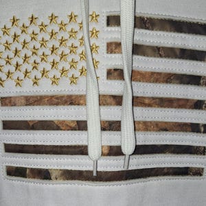 May include: White hoodie featuring an American flag design. The flag incorporates gold embroidered stars and camouflage stripes. White drawstrings are visible. The hoodie is a patriotic apparel item.