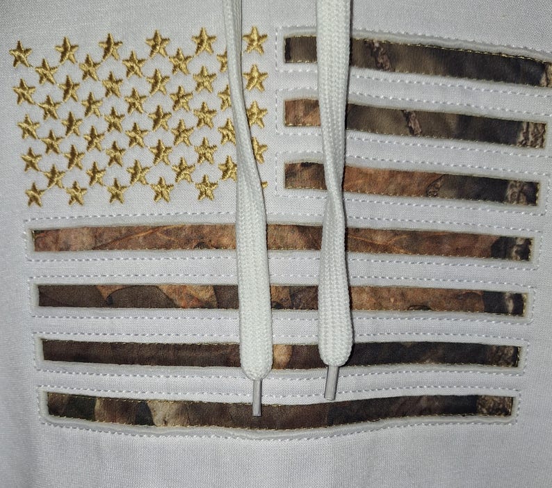 May include: White hoodie featuring an American flag design. The flag incorporates gold embroidered stars and camouflage stripes. White drawstrings are visible. The hoodie is a patriotic apparel item.