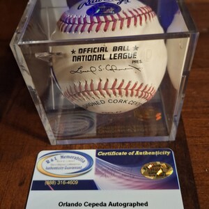 Op de afbeelding: Een witte honkbal gesigneerd door Orlando Cepeda, een voormalige Major League Baseball-speler. De bal bevindt zich in een transparante kunststof display met een certificaat van echtheid. De bal is een offici&euml;le National League-bal en heeft het Rawlings-logo erop. Het certificaat van echtheid is van R&L Memorabilia.
