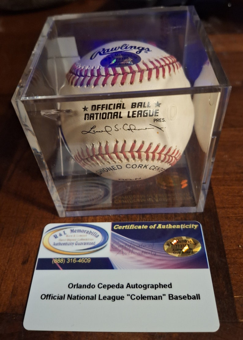 Op de afbeelding: Een witte honkbal gesigneerd door Orlando Cepeda, een voormalige Major League Baseball-speler. De bal bevindt zich in een transparante kunststof display met een certificaat van echtheid. De bal is een offici&euml;le National League-bal en heeft het Rawlings-logo erop. Het certificaat van echtheid is van R&L Memorabilia.