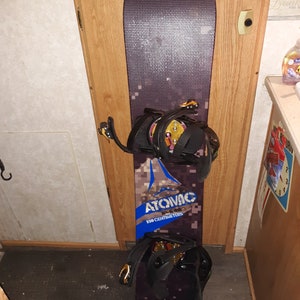 May include: A black and brown snowboard with a blue and white logo that says "Atomic 160 centimeters". The snowboard has two black bindings with gold buckles.