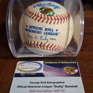Op de afbeelding: Een witte honkbal met rode naden en de tekst "OFFICIAL BALL AMERICAN LEAGUE" en "Hana A. Budig PRES." in zwart. De bal bevindt zich in een doorzichtige plastic behuizing met een echtheidscertificaat van R&L Memorabilia.