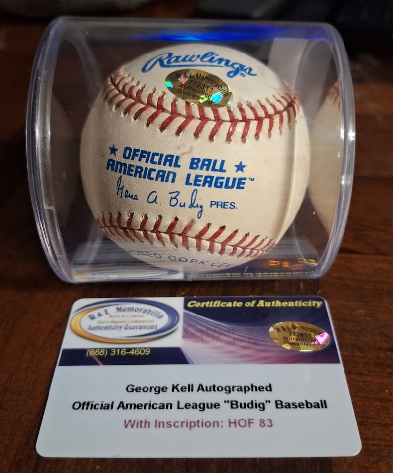 Op de afbeelding: Een witte honkbal met rode naden en de tekst "OFFICIAL BALL AMERICAN LEAGUE" en "Hana A. Budig PRES." in zwart. De bal bevindt zich in een doorzichtige plastic behuizing met een echtheidscertificaat van R&L Memorabilia.