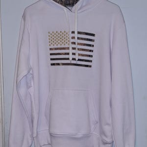 May include: White hooded sweatshirt featuring a camouflage-patterned hood lining. The front displays an American flag design with gold stars and camouflage stripes. The sweatshirt has a front pocket and drawstring hood.