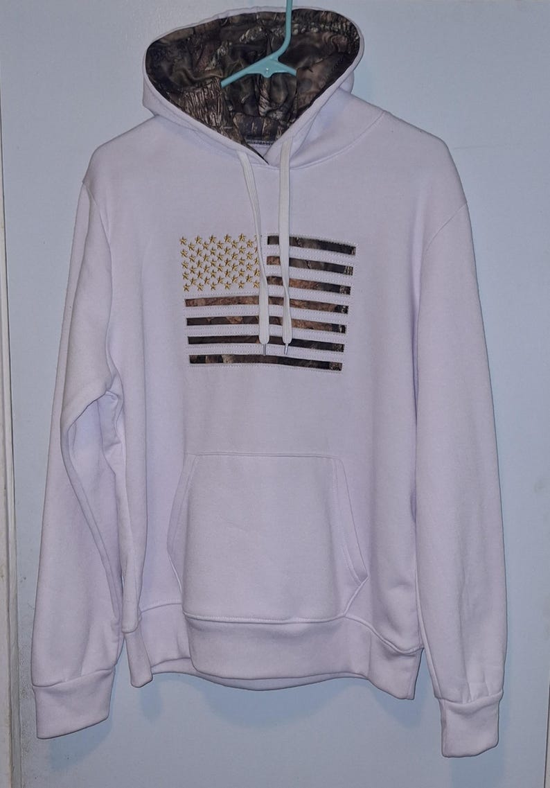 May include: White hooded sweatshirt featuring a camouflage-patterned hood lining. The front displays an American flag design with gold stars and camouflage stripes. The sweatshirt has a front pocket and drawstring hood.