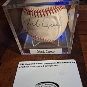 Op de afbeelding: Een witte honkbal gesigneerd door Orlando Cepeda in een transparante vitrine met een witte kaart waarop staat: "R&L Memorabilia Inc. garandeert de authenticiteit van al onze handgesigneerde handtekeningen." en een zwart-wit logo met de tekst "R&L Memorabilia Echtheitsgarantie".