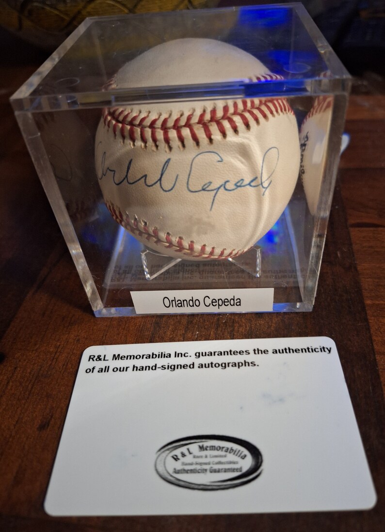 Op de afbeelding: Een witte honkbal gesigneerd door Orlando Cepeda in een transparante vitrine met een witte kaart waarop staat: "R&L Memorabilia Inc. garandeert de authenticiteit van al onze handgesigneerde handtekeningen." en een zwart-wit logo met de tekst "R&L Memorabilia Echtheitsgarantie".