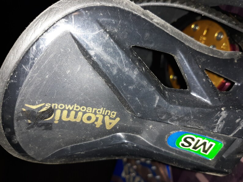 May include: Close-up of a black snowboard binding with a gold "Atom! snowboarding" logo and a green and blue "MS" sticker.