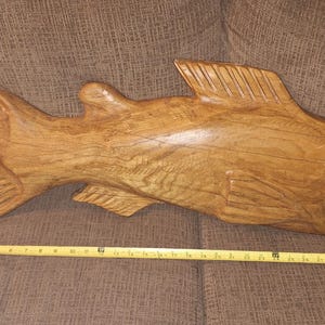 May include: A wooden carving of a fish with a detailed tail and scales. The fish is painted in a light brown color and has a realistic appearance.