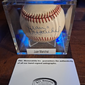 Op de afbeelding: Een witte honkbal gesigneerd door Juan Marichal, tentoongesteld in een heldere acryl vitrine met een blauwe lichtbasis. De vitrine heeft een wit label met de tekst "R&L Memorabilia Inc. garandeert de authenticiteit van al onze handgesigneerde handtekeningen." en een zwart-wit logo met de tekst "R&L Memorabilia Echtheid gegarandeerd".