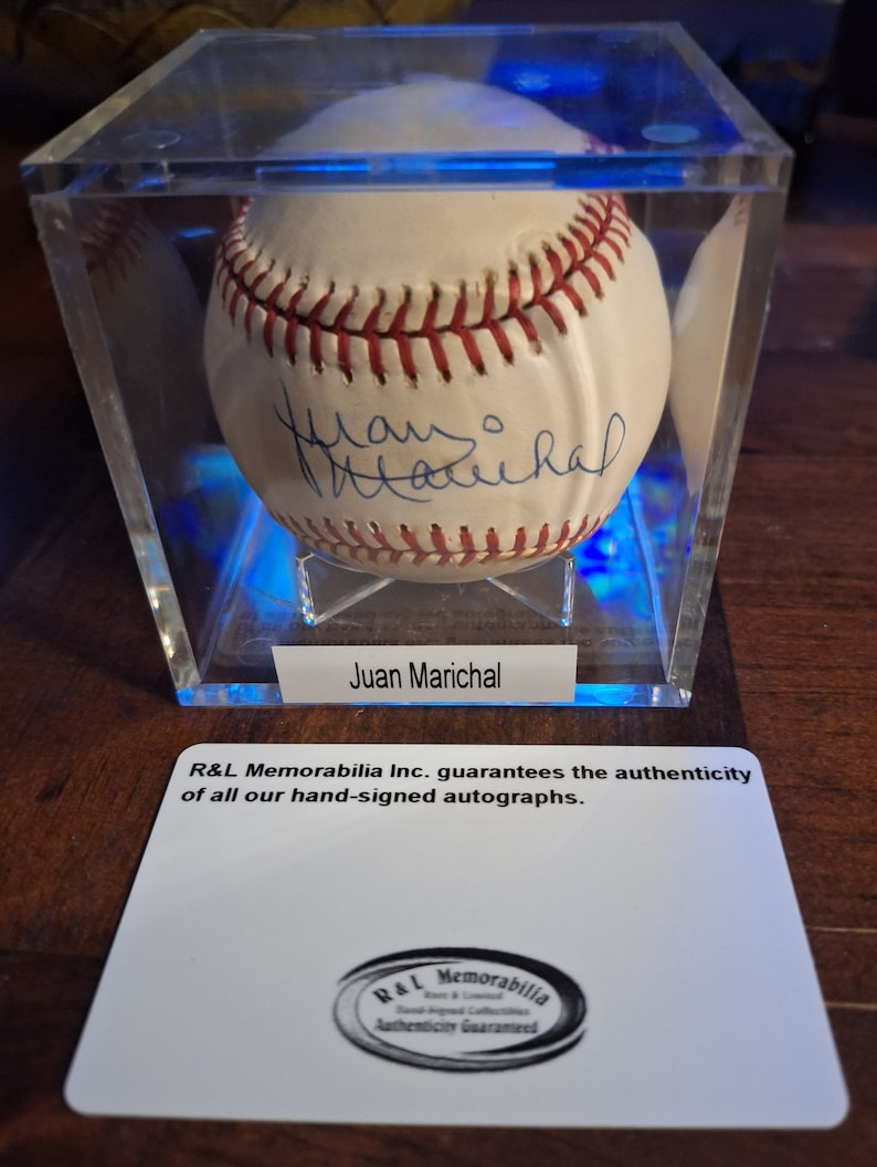 Op de afbeelding: Een witte honkbal gesigneerd door Juan Marichal, tentoongesteld in een heldere acryl vitrine met een blauwe lichtbasis. De vitrine heeft een wit label met de tekst "R&L Memorabilia Inc. garandeert de authenticiteit van al onze handgesigneerde handtekeningen." en een zwart-wit logo met de tekst "R&L Memorabilia Echtheid gegarandeerd".