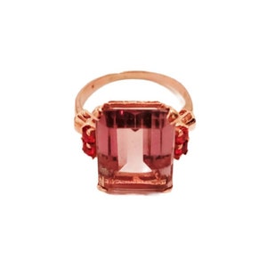 May include: A rose gold ring with a large, rectangular, purple gemstone. The gemstone is surrounded by smaller, red gemstones.