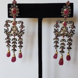 May include: A pair of dangle earrings with a gold and silver design. The earrings feature red ruby stones and are accented with smaller diamonds. The earrings are suspended from a small ruby stone.