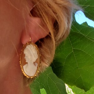 May include: A pair of gold cameo earrings with a white cameo of a woman's profile. The earrings are hanging from a person's ear.