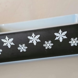 Puede incluir: Fuente de horno rectangular negra con diseños de copos de nieve blancos. El interior es azul claro y el borde es blanco. El plato tiene un asa en cada extremo. Un artículo de cocina festivo.