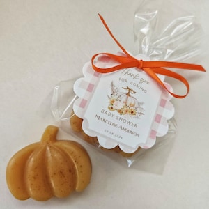 May include: A small, orange pumpkin-shaped soap bar with a thank you tag. The tag has a pink and white checkered background with a white pumpkin and floral design. The text on the tag reads "Thank you for coming, Baby Shower Marceline Anderson 09.08.2024".