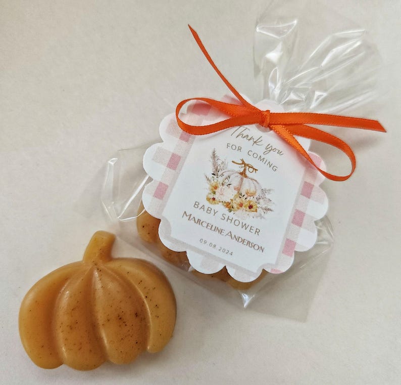May include: A small, orange pumpkin-shaped soap bar with a thank you tag. The tag has a pink and white checkered background with a white pumpkin and floral design. The text on the tag reads "Thank you for coming, Baby Shower Marceline Anderson 09.08.2024".