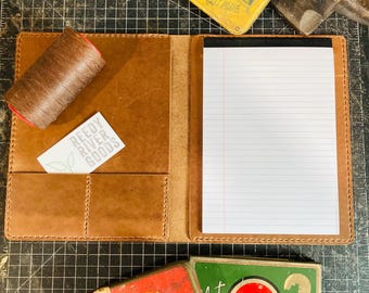 May include: A brown leather notebook with a notepad and pockets, a spool of brown thread, and a business card. Vintage metal tins with text are scattered around the notebook. An anvil is in the background.