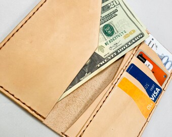 May include: A light tan leather wallet, open to display a twenty-dollar bill and several credit cards. The wallet has multiple card slots and a cash pocket. The edges are stitched with a contrasting thread, and the leather has a natural, unfinished look.