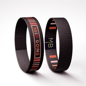 Può includere: Due braccialetti neri intrecciati con strisce colorate e testo. Un braccialetto dice "KEEP GOING" e l'altro dice "MB".