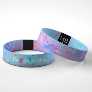 Puede incluir: Dos pulseras azul claro con diseños de salpicaduras de pintura coloridas y las palabras "Be Artistic". Una pulsera tiene un cierre de velcro negro. La otra pulsera tiene "MB" impreso en una etiqueta negra.