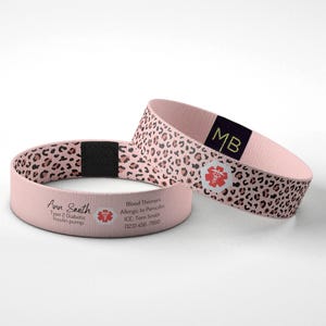 May include: Two pink medical alert bracelets. One has a leopard print design, the other has the name "Ann Smith" and medical information printed on it. Both have a red medical symbol.