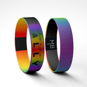 Puede incluir: Dos pulseras tejidas arcoíris con texto negro. Una pulsera dice "Ally" y la otra dice "MB".