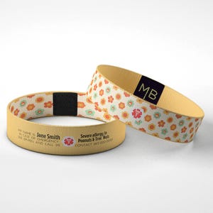 May include: Two beige medical alert wristbands. One band features a floral pattern with orange, pink, and green flowers. The other band has text that reads "MY NAME Jane Smith, RELEASE OF EMERGENCY, USE ER PEN AND CALL 98".
