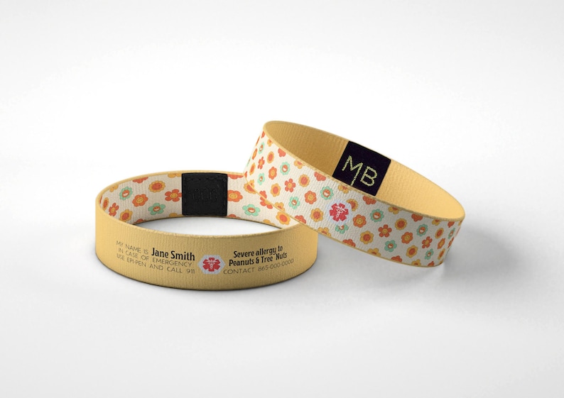 May include: Two beige medical alert wristbands. One band features a floral pattern with orange, pink, and green flowers. The other band has text that reads "MY NAME Jane Smith, RELEASE OF EMERGENCY, USE ER PEN AND CALL 98".