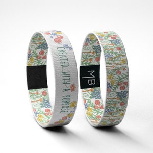 May include: Two white wristbands with a floral pattern and the text "CREATED WITH A PURPOSE" on one of the bands. The bands have a black adjustable closure.