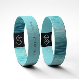 May include: Two light blue wristbands with a watercolor effect. One band has the text "REMEMBER TO BREATHE" printed in gray. Each band has a black tag with a geometric design. The bands are made of a flexible material.